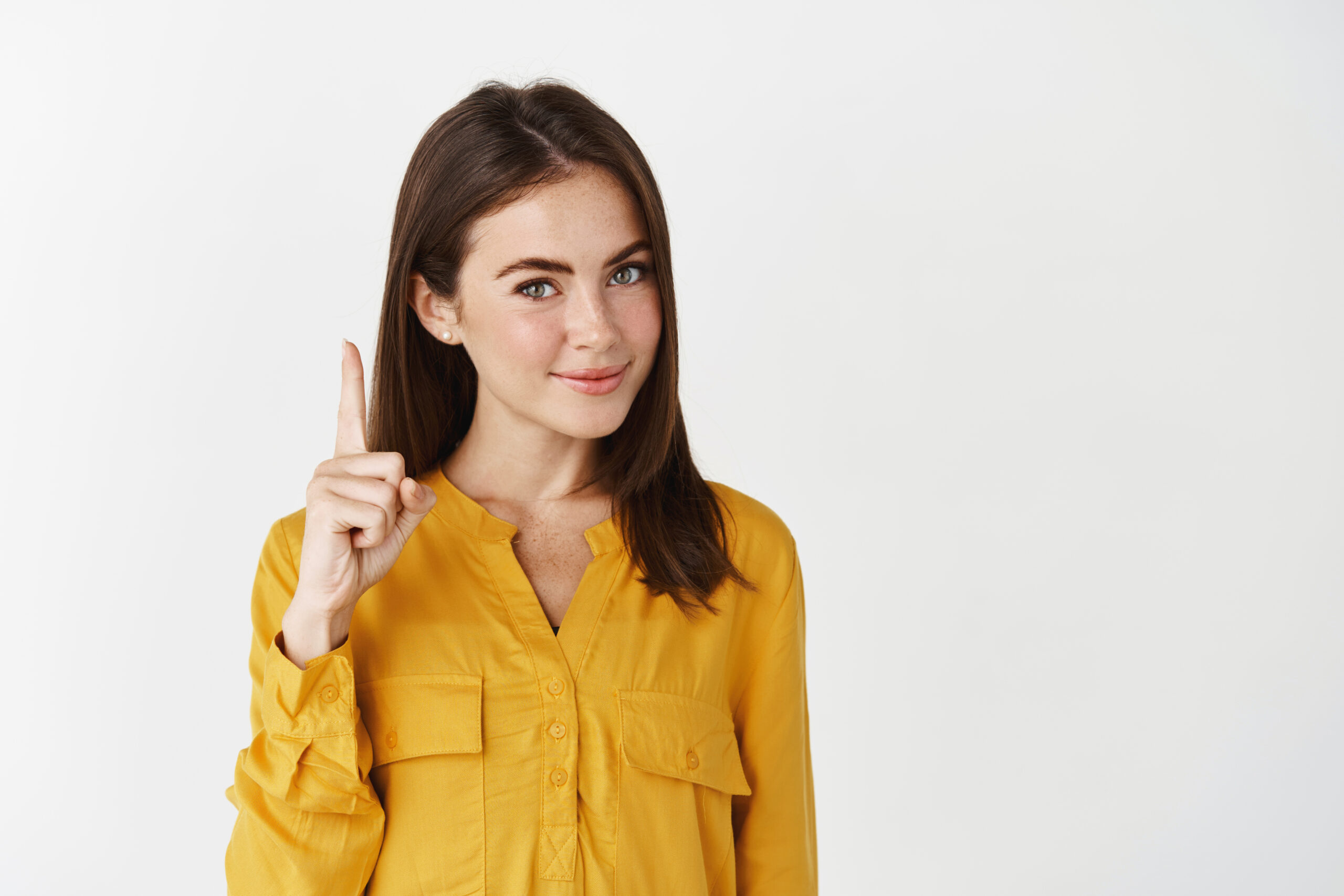 Image of young thoughtful woman showing advertisement, pointing finger up and smiling at camera, giving knowing look, standing over white background
